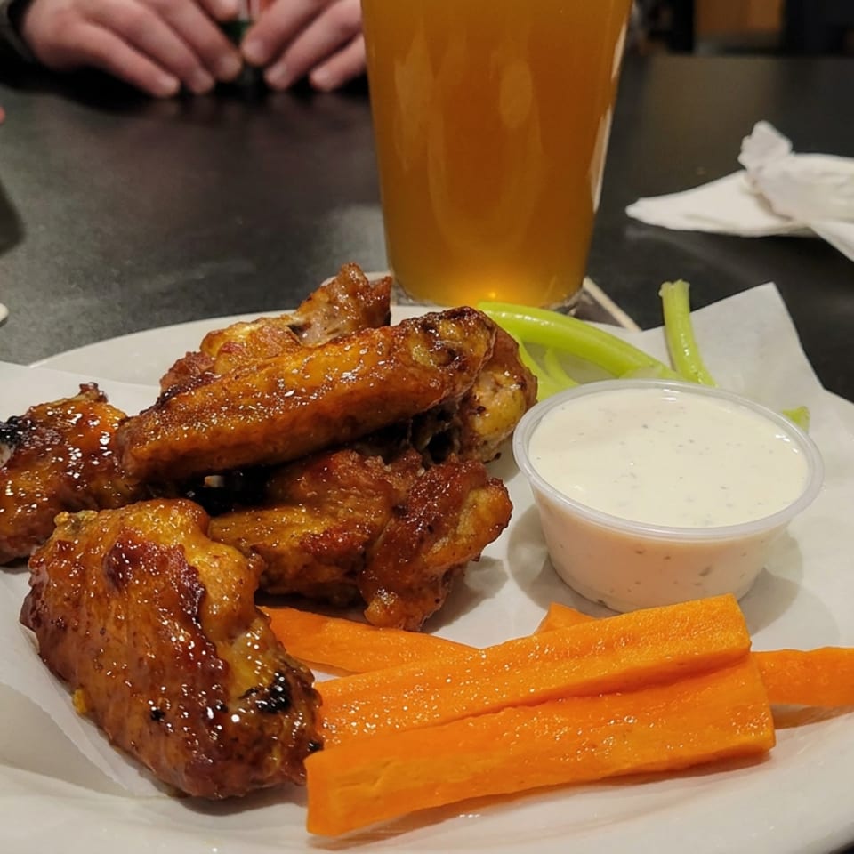 Glazed chicken wings with carrot and celery sticks, ranch dipping sauce, and beer on a restaurant table.