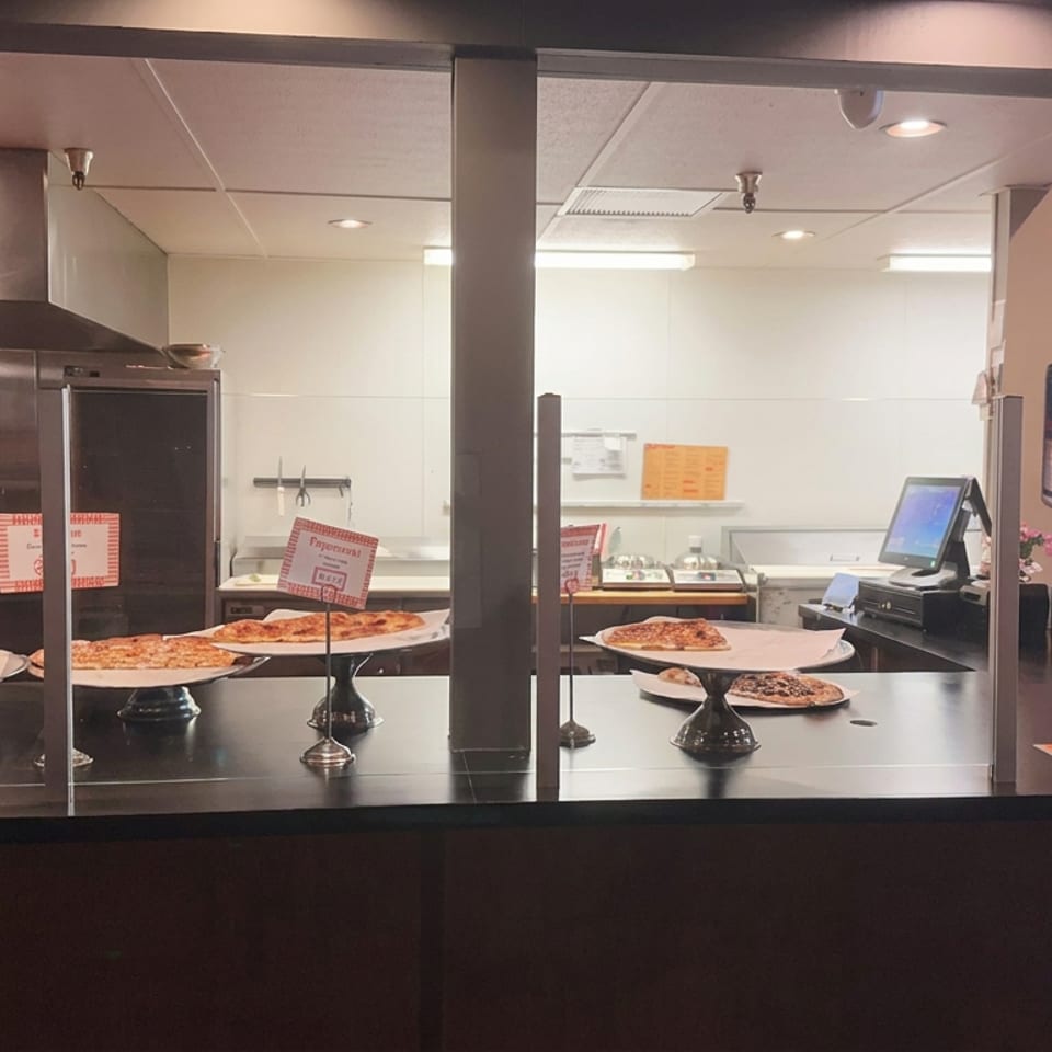 Restaurant counter displaying assorted pizza slices on elevated serving stands, with an open kitchen, register, and colorful wall mural.