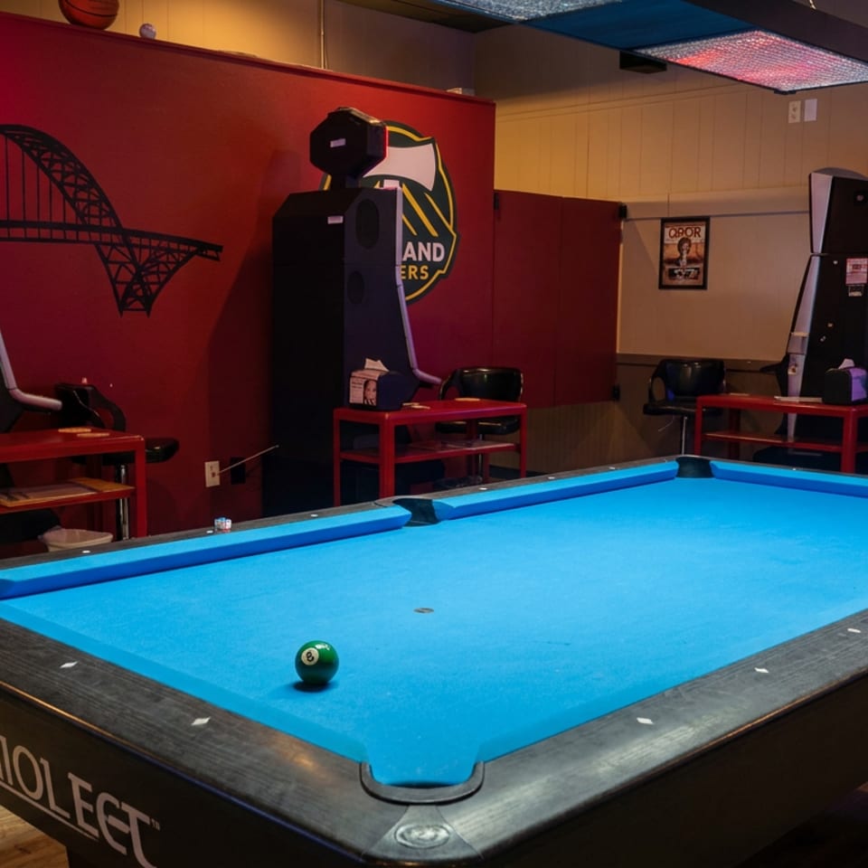 Pool table with blue felt and billiard ball in a sports bar featuring arcade basketball games, televisions, and red walls.